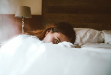 Brunette female lying in bed, peering out from behing the duvet, looking anxious