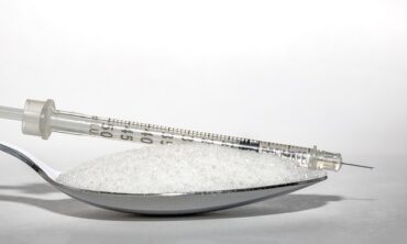 Close up of a teaspoon filled with sugar and in insulin injection is lying on top