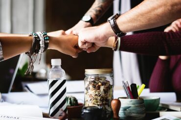 Four arms reaching out to fist bump over a desk with various bits and bobs on it - indicating teamwork and positivity