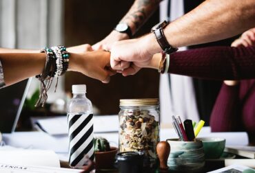 Four arms reaching out to fist bump over a desk with various bits and bobs on it - indicating teamwork and positivity