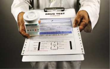 Close u up for a medical drug test form on a clipboard being held by a doctor who is also holding a jar of pills