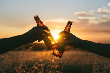 silhouette of two hands holding two beer bottles that are being touched together in cheers. Background is a bright orange sunset.