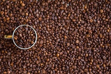 Background filled with coffee beans and a single cup is sat on top on the left hadn side, also filled with coffee beans