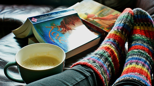 close up of a womans feet in brightly coloured striped woolen socks on a bed with two open books and a cup of coffee next to her.