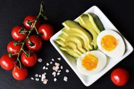 Examples of keto foods laid out on a white square plate on a black background. Includes vine tomatoes, cliced avocado and halved hard boiled egg