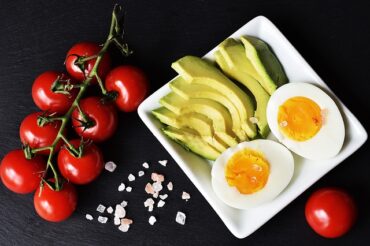 Examples of keto foods laid out on a white square plate on a black background. Includes vine tomatoes, cliced avocado and halved hard boiled egg