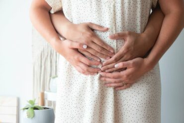 Mid section of a prgenant lady weaing a white shirt dress holding. Her partner is stood behing her and their hand are resting over the pregnancy bump.