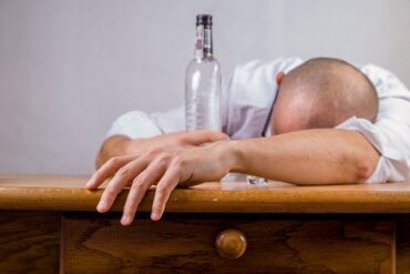 Man lat down leaning over a table with hid bald head restin gon his arms and hugging an empty bottle glass bottle. Suggesting he has drunk the alcoholic substance inside and is an alcoholic who needs help.