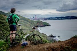 Male cyclist wearing black shorts and a green long sleeved top stood next to his bike looking out across a lake