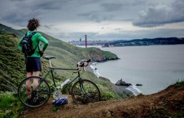 Male cyclist wearing black shorts and a green long sleeved top stood next to his bike looking out across a lake
