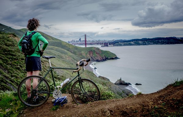 Male cyclist wearing black shorts and a green long sleeved top stood next to his bike looking out across a lake