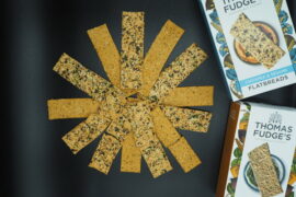 Thomas Fudge's Flatbread laid out in a circle fanning out from the middle. The background is black and the packaging is placed to the right hand side.