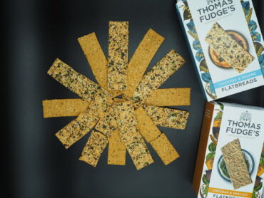 Thomas Fudge's Flatbread laid out in a circle fanning out from the middle. The background is black and the packaging is placed to the right hand side.