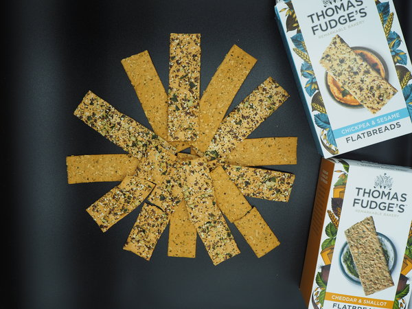 Thomas Fudge's Flatbread laid out in a circle fanning out from the middle. The background is black and the packaging is placed to the right hand side.