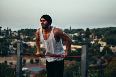 Man in his twenties wearing a white vest, jeans and a dark beanie hat is using outdoor gym equipment. He is pushing himself up on a metal bar to show how calisthenics is good for strength training. In the background there is a suburban town.