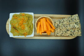 white square bowl of carrot and coriander hummus topped with grated carrot. next to it is a round wooden bowol filled with carrot sticks and three seeded flatbreads. they are lined up on a wooden flat plate and set against a black background