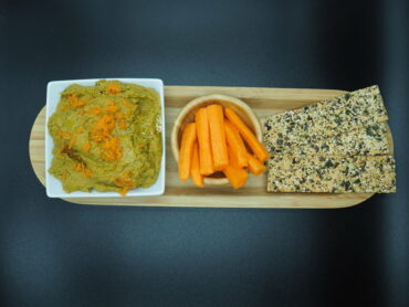 white square bowl of carrot and coriander hummus topped with grated carrot. next to it is a round wooden bowol filled with carrot sticks and three seeded flatbreads. they are lined up on a wooden flat plate and set against a black background