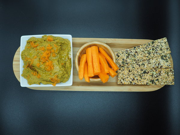 white square bowl of carrot and coriander hummus topped with grated carrot. next to it is a round wooden bowol filled with carrot sticks and three seeded flatbreads. they are lined up on a wooden flat plate and set against a black background