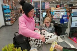 Mum and daughter doing a healthy family food shop at Marks and Spencer. They are at the till putting some bananas in a carrier bag. Mum is wearing a black bobble hat and a pink jumper and is smiling. Daughter is wearing a silver puffa jacket and is wearing glasses.