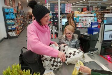 Mum and daughter doing a healthy family food shop at Marks and Spencer. They are at the till putting some bananas in a carrier bag. Mum is wearing a black bobble hat and a pink jumper and is smiling. Daughter is wearing a silver puffa jacket and is wearing glasses.