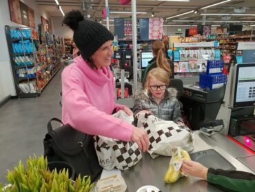 Mum and daughter doing a healthy family food shop at Marks and Spencer. They are at the till putting some bananas in a carrier bag. Mum is wearing a black bobble hat and a pink jumper and is smiling. Daughter is wearing a silver puffa jacket and is wearing glasses.