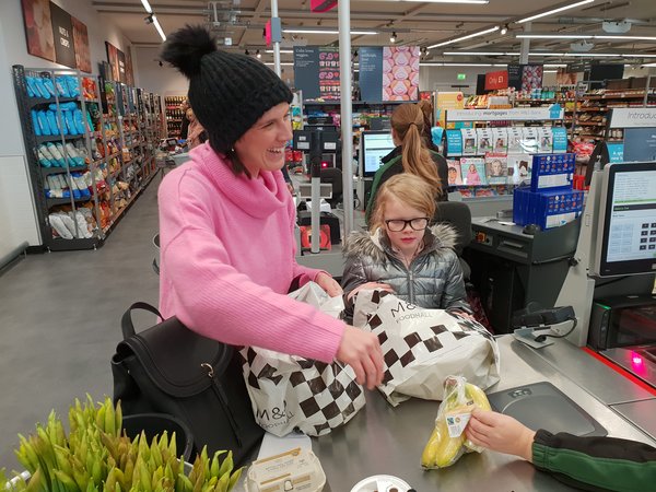 Mum and daughter doing a healthy family food shop at Marks and Spencer. They are at the till putting some bananas in a carrier bag. Mum is wearing a black bobble hat and a pink jumper and is smiling. Daughter is wearing a silver puffa jacket and is wearing glasses.