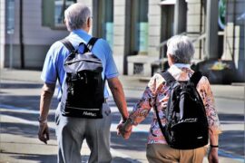 an older coupl holding hands stood with their backs to the camera. They both have black rucksakcs and look as though they are exploring a foreign city together in retirement.