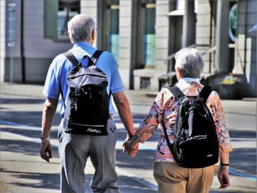 an older coupl holding hands stood with their backs to the camera. They both have black rucksakcs and look as though they are exploring a foreign city together in retirement.