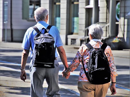 an older coupl holding hands stood with their backs to the camera. They both have black rucksakcs and look as though they are exploring a foreign city together in retirement.