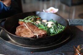 Blac k frying pan containing a t-bone steak and green vegetables