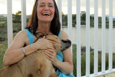 Middle aged woman sat cuddling her dog and laughing. She looks younger than she is and really happy and full of life. She is wearing a bright blue sleeveless dress and leaning against some white railings which look out over a lake.