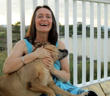 Middle aged woman sat cuddling her dog and laughing. She looks younger than she is and really happy and full of life. She is wearing a bright blue sleeveless dress and leaning against some white railings which look out over a lake.
