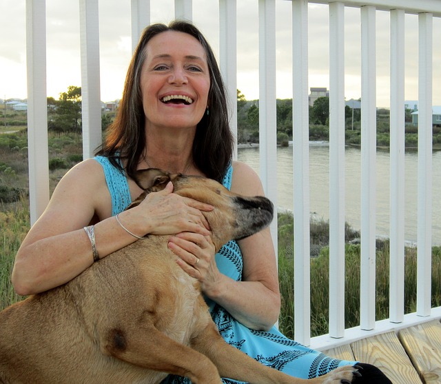 Middle aged woman sat cuddling her dog and laughing. She looks younger than she is and really happy and full of life. She is wearing a bright blue sleeveless dress and leaning against some white railings which look out over a lake.