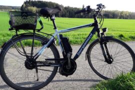 A black and white e-bike stood up in the middle of a country road. There is a green field and woodland in the background and it is a sunny day, perfect for cycling to work.