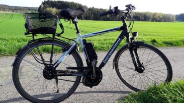 A black and white e-bike stood up in the middle of a country road. There is a green field and woodland in the background and it is a sunny day, perfect for cycling to work.
