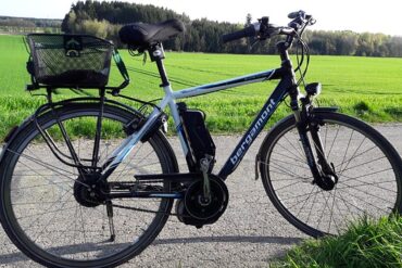A black and white e-bike stood up in the middle of a country road. There is a green field and woodland in the background and it is a sunny day, perfect for cycling to work.