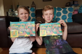 2 children, one girl and one boy sat in a living room holding up 2 boxes of Munchachos snack boxes for kids