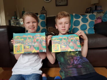 2 children, one girl and one boy sat in a living room holding up 2 boxes of Munchachos snack boxes for kids
