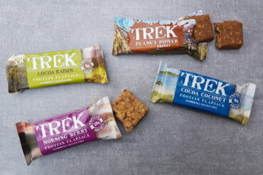A selection of 4 Trek protein flapjacks and protein energy bars laid out on a light grey slate background. 2 packets are open revealing the bars and the other two are closed.
