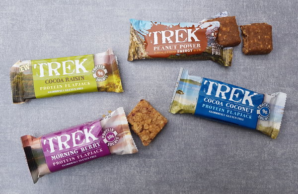 A selection of 4 Trek protein flapjacks and protein energy bars laid out on a light grey slate background. 2 packets are open revealing the bars and the other two are closed.