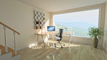 A light airy room with a massive wqindow overlooking a beautiful vista. The room has light wooden floors and cream walls and there is a corner desk with a computer on it and a chair. On the wall is a piece of angular white wall art. It looks like a very unclutteered tranquil space.