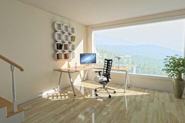 A light airy room with a massive wqindow overlooking a beautiful vista. The room has light wooden floors and cream walls and there is a corner desk with a computer on it and a chair. On the wall is a piece of angular white wall art. It looks like a very unclutteered tranquil space.
