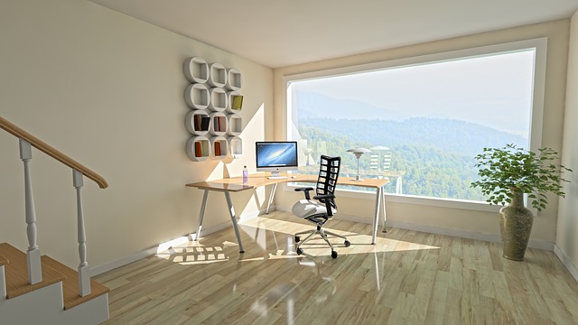 A light airy room with a massive wqindow overlooking a beautiful vista. The room has light wooden floors and cream walls and there is a corner desk with a computer on it and a chair. On the wall is a piece of angular white wall art. It looks like a very unclutteered tranquil space.