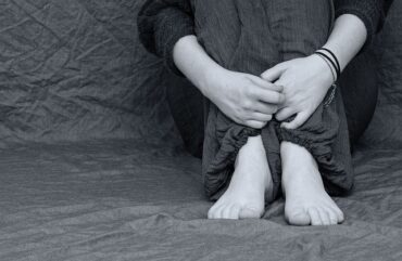 black and white image of someones hand feet and knees sat scrunched up clutching themselves