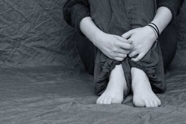 black and white image of someones hand feet and knees sat scrunched up clutching themselves