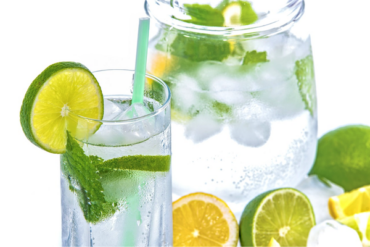 glass filled with clean filtered water with a mint leaf, ice cubes a green straw and a slice of lime on the side. There is a clear glass jug filled with the same contents in the background and there is hald a sliced lemon and lime next to the glass.