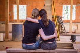 Two femasle friends sat side by side with their backs to the camera. they ahve their arms around one another to help support and show their friendship. They are in what looks like a garage or work room.