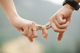 close up of a male and female hand joined by interlocking middle fingers. the woman is wearing a ring and the man has on a wrist watch. On their middle fingers they both had small tattoos of an achor showing they work hard at keeping their relationship strong.