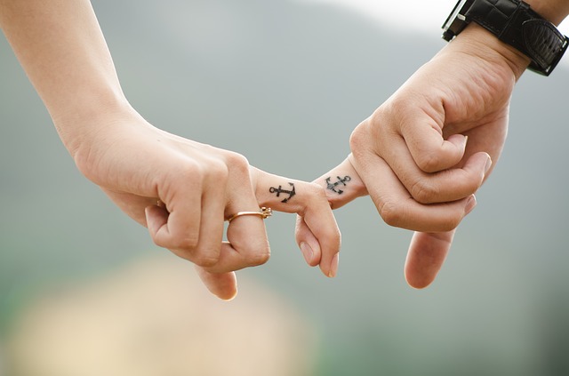 close up of a male and female hand joined by interlocking middle fingers. the woman is wearing a ring and the man has on a wrist watch. On their middle fingers they both had small tattoos of an achor showing they work hard at keeping their relationship strong.