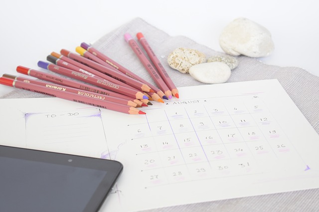 a white calendar sheet laid out on a desk with some coloured pencils the corner of a tablet device and some pebbles